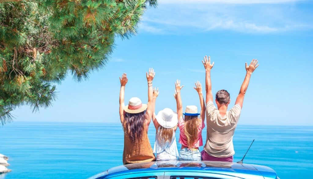 persone sedute sul tettuccio della macchina con le braccia al cielo davanti al mare 